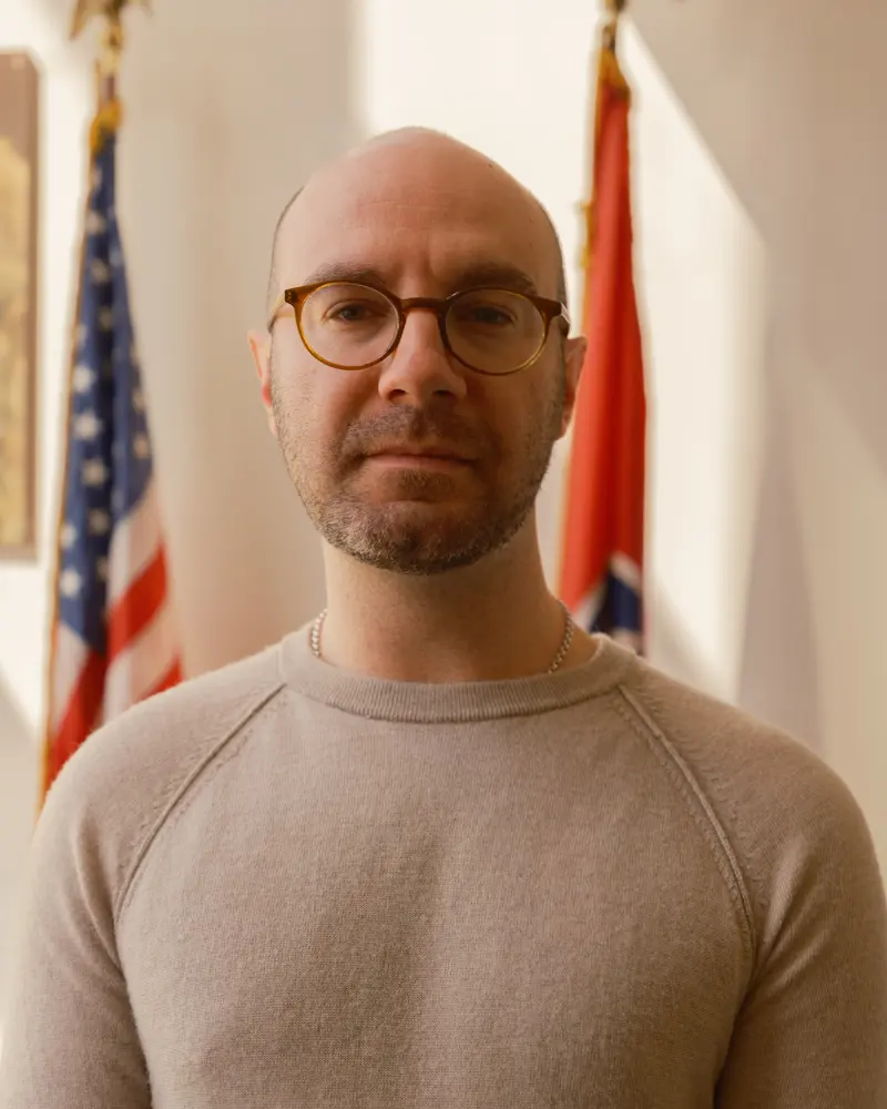 A white man with round, brown glasses and short beard stands in front of a U.S. and a Tennessee flag.