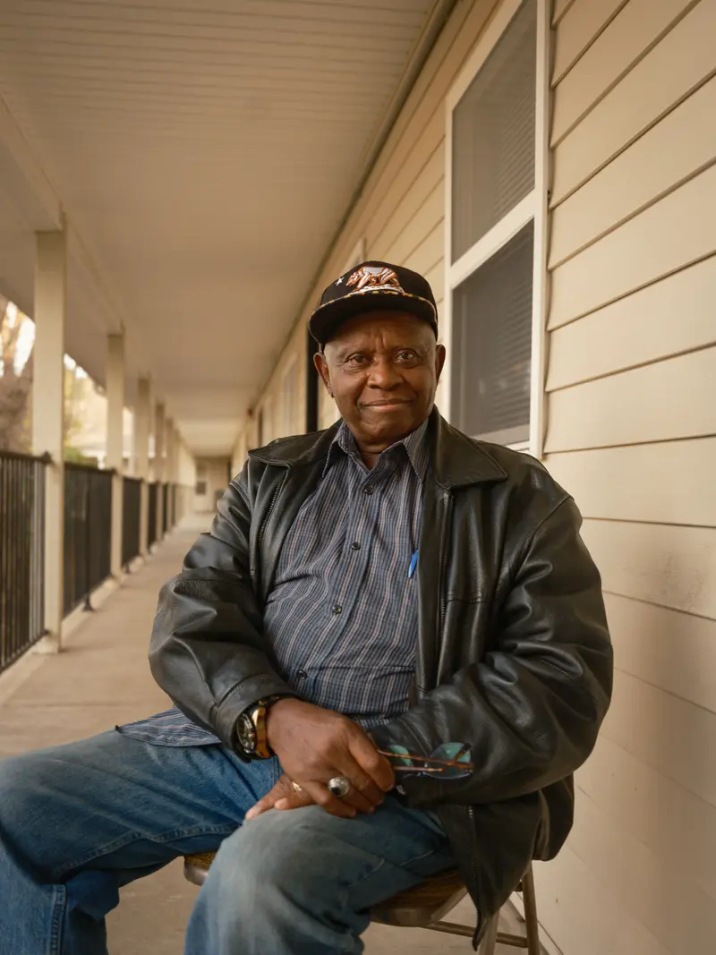 A Black man in jeans and a leather jacket sits on a folding chair on a long outdoor walkway with metal railings.