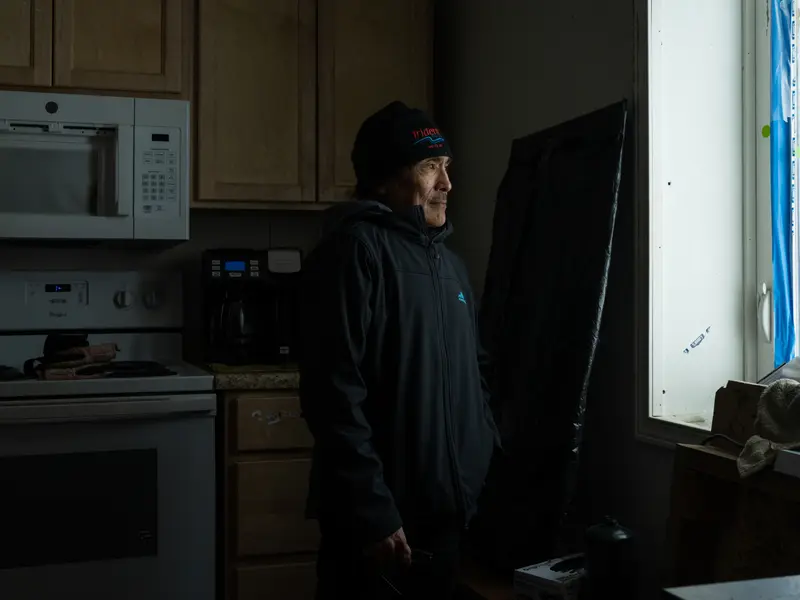A man wearing a black hoodie and black hat stands in a dark kitchen, looking out a lit window.