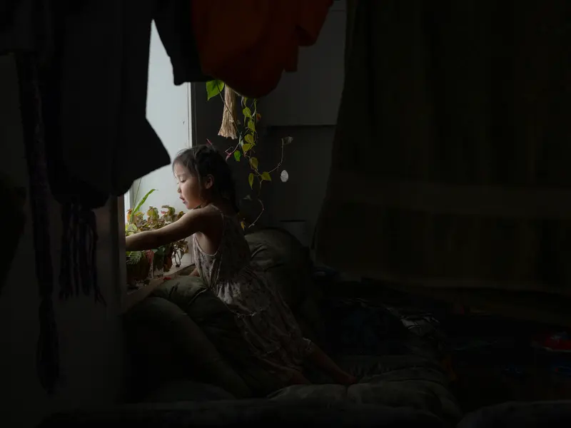 A little girl stands at a window near plants while the rest of the room is in darkness.
