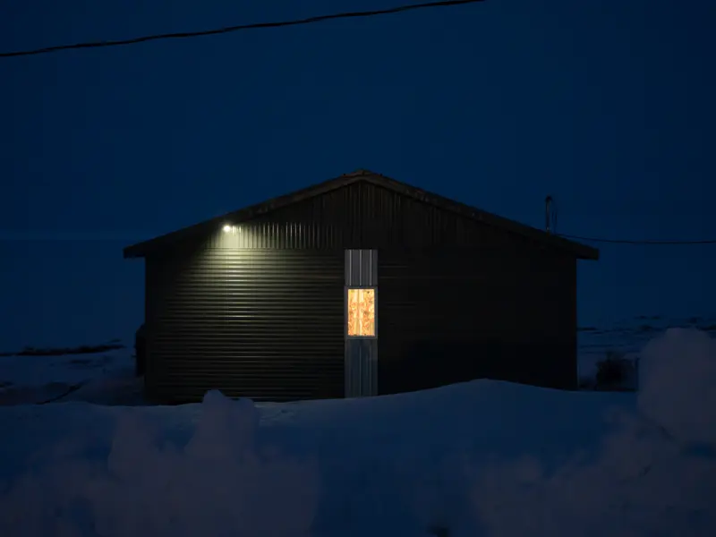 A nearly black one-story house with one lit window and a roof light sits in snow surrounded by blue-tinged darkness.