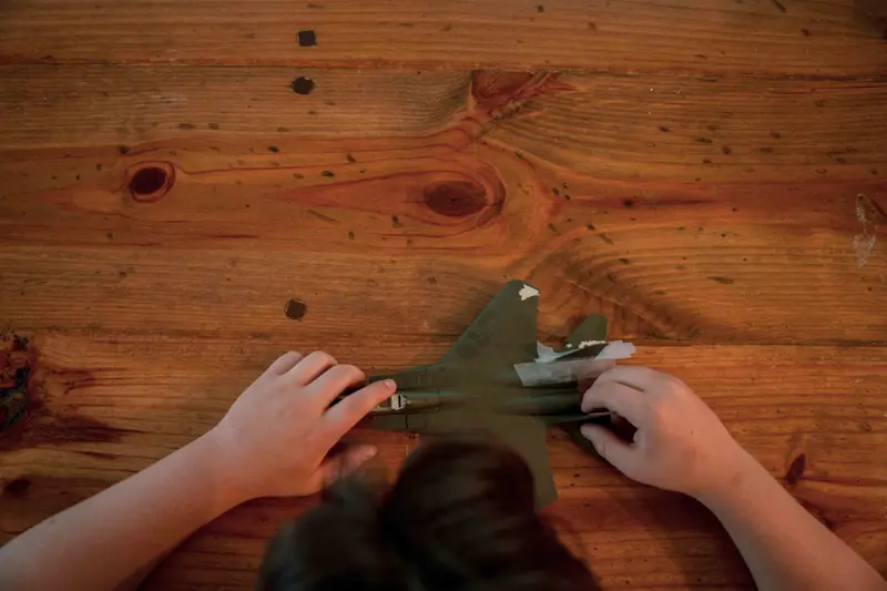 An aerial view of a boy’s hands touching a model fighter jet on a wooden table.
