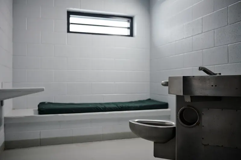 A white cinderblock room with a narrow window, a thin cot on a cinder block base, a small shelf and a metal toilet.