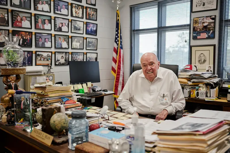 Bean sits at his desk smiling. The desk and room are cluttered with photos of Bean posing with other people, memorabilia including Trump and Obama figurines, file folders and an American flag.