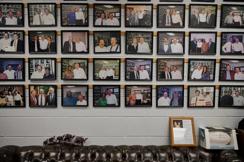 A grid of thirty-two portraits of Bean posing with different people hang on a cinder block wall.