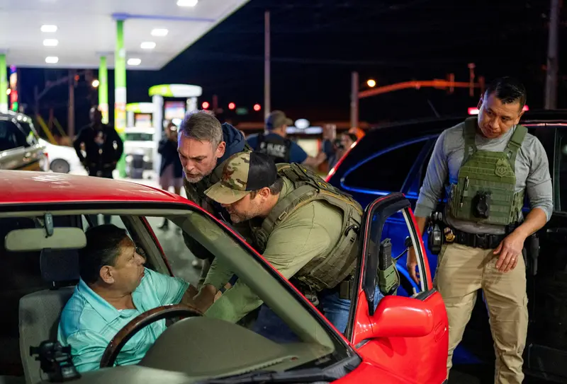 Two officers in armored vests attempt to pull a man out of the driver’s seat of a car. A third officer in a vest stands behind them, and several more are visible in the background.