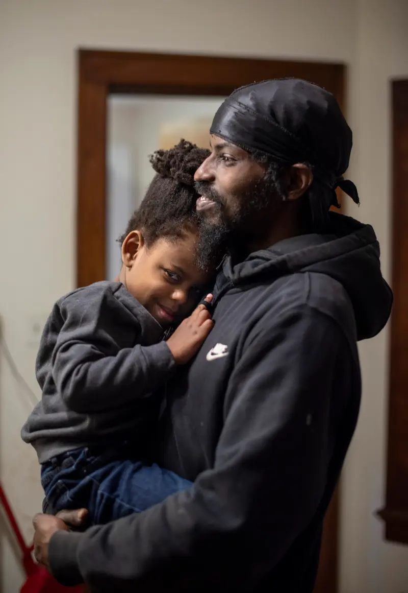 Moses holds Samir to his chest as Moses smiles and Samir smiles shyly at the camera with his finger wrapped around the drawstring of Moses’ hoodie.