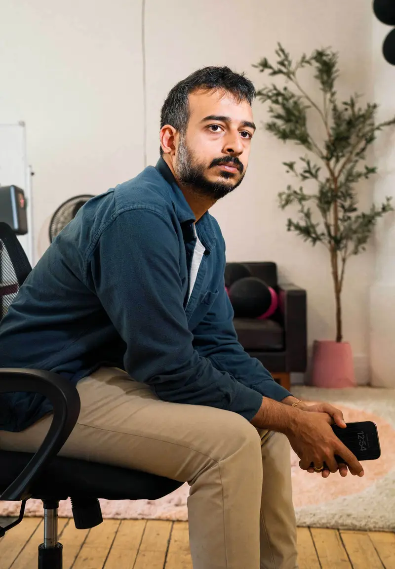 A man wearing khakis and a blue jacket sits on a swivel chair in an office in a renovated loft while holding a smartphone.