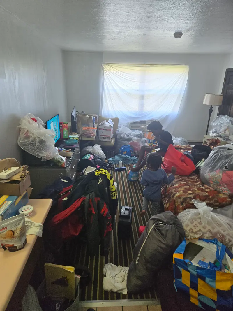Taylor and Samir watch television in the motel room filled with suitcases and bags. A bedsheet acts as a curtain for the window.