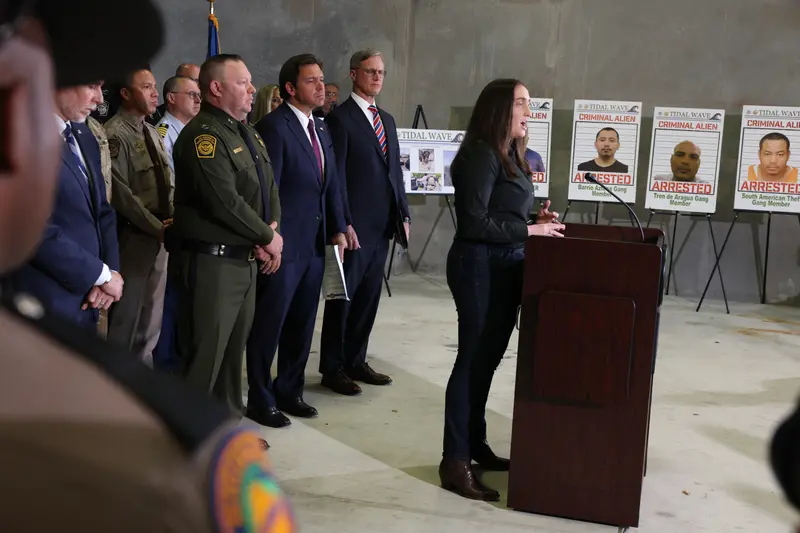 Rows of men in uniforms and suits stand behind a woman speaking at a lectern. Large photographs of mugshots are propped up on easels in the background.