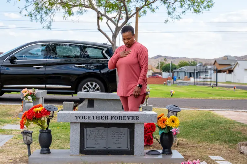 A photo of a woman wearing a pink outfit, standing with one hand over her chest, behind a gravestone inscribed with Bible verses and the words “together forever” in a grassy cemetery with a black car and tree in the background.