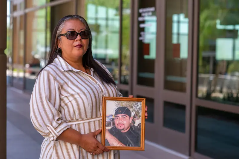 A woman wearing a tan and white dress and sunglasses holds a photo of a man in a black shirt and black hat, in front of the glass facade of a building.