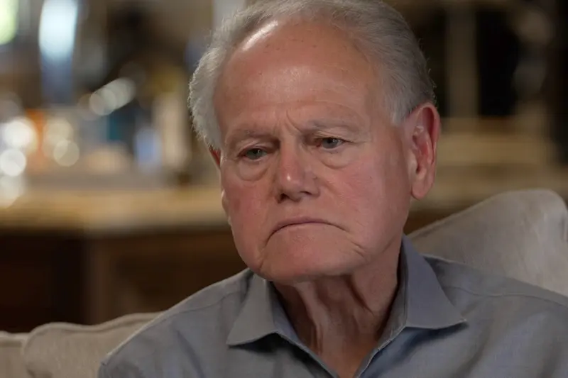 A close portrait of a man with grey hair and a collared dark grey shirt seated on a tan couch in a nondescript room.
