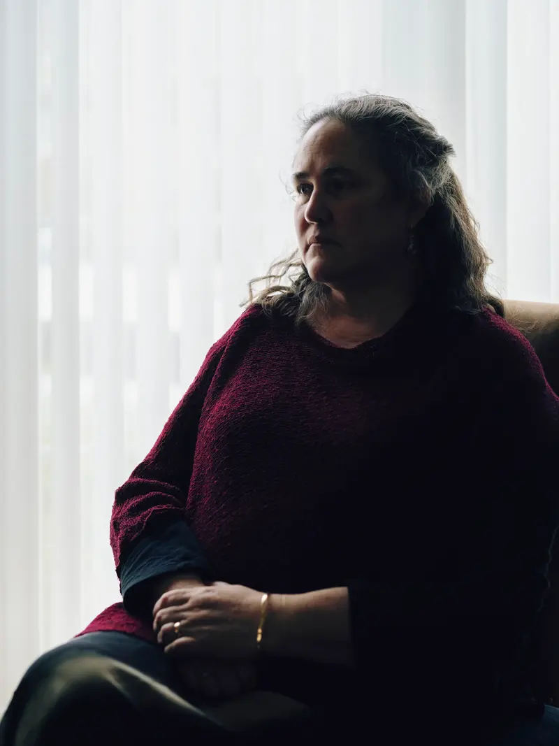A woman with shoulder-length hair sits in a chair, her face half in shadow, with blinds behind her.