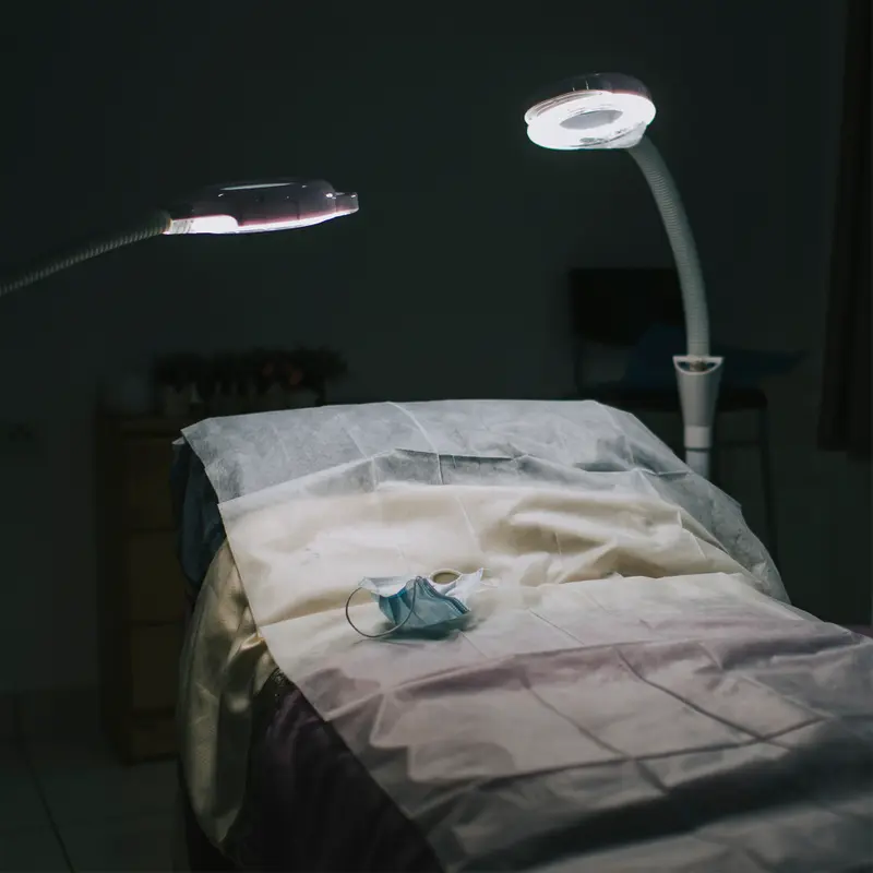 A blue paper mask rests on an empty medical exam bed illuminated by two bright lights in a dark room.