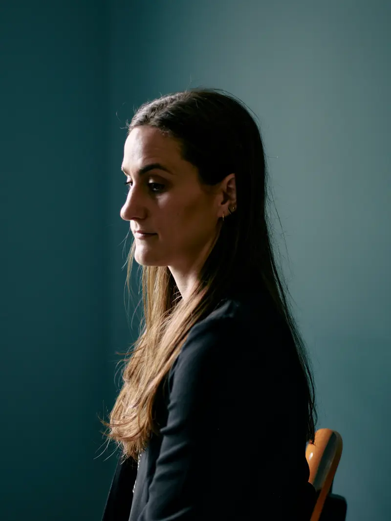 A woman with long brown hair looks solemnly down, seated in a chair with light falling on half of her face.