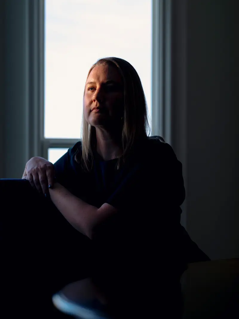 A woman sitting in a dark room, mostly in shadow, with a pocket of light falling on her face.
