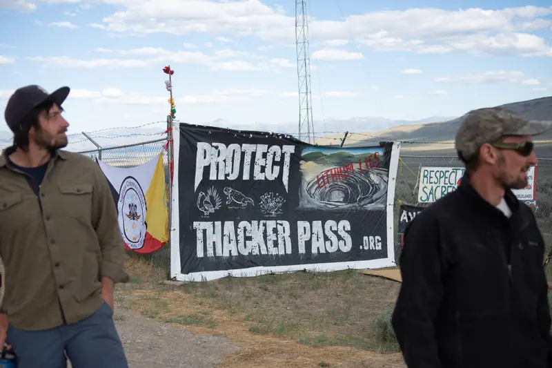 Two men stand in front of a chain-link fence, where a sign is hung that says “Protect Thacker Pass” and “Lithium Lies.”