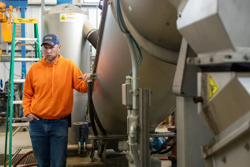 A man in jeans and a bright orange sweatshirt and stands next to a metal pipe that’s several feet in diameter.