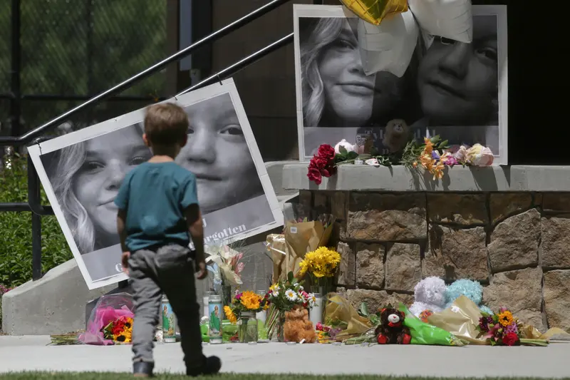 A small boy looks at two oversize black-and-white photos of children attached to a railing. Beneath the photos are teddy bears, flower bouquets and votive candles.
