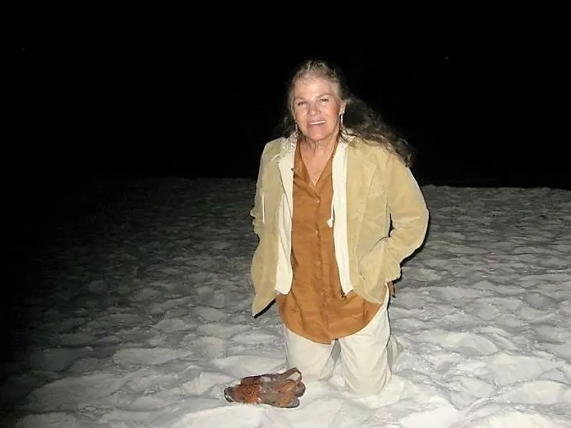 A woman with long dark hair smiles, kneeling in white sand with darkness surrounding her.