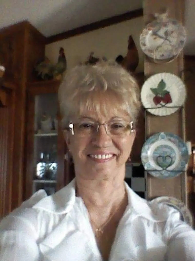An older woman with short, light-colored hair smiles in front of a wall of decorative plates and other knickknacks.