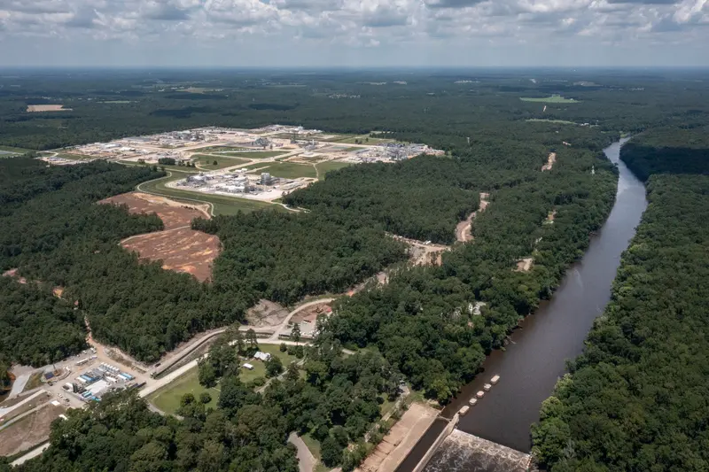 A bird’s-eye view of a river winding through a dense forest near a large industrial site, with rolling clouds above.