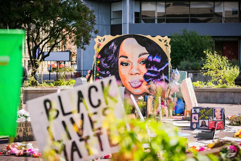 A memorial in a public square includes a painted portrait of Breonna Taylor, a Black Lives Matter sign and a handwritten sign reading, “She was asleep.”
