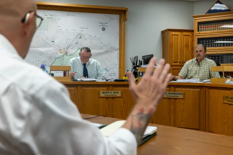 A man in a white shirt and glasses sits at a wooden table with his back to the camera and his hand outstretched towards two men seated facing him at a row of wooden desks.