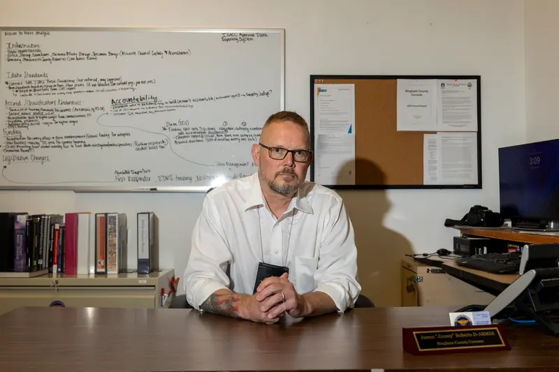 A man wearing a long-sleeve white button-down shirt and glasses sits at a wooden desk looking directly at the camera. Behind him is a white board covered in writing, a bulletin board and a bookcase.