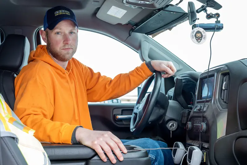 A man wearing a navy baseball cap and neon orange sweatshirt sits inside a car with one hand on the steering wheel, looking toward the camera.