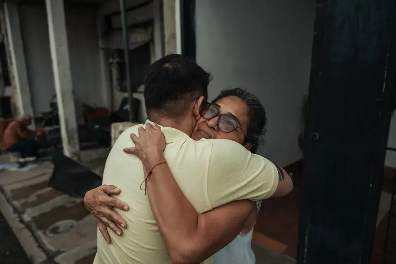 Eine Frau umarmt einen Mann in einem gelben Hemd fest, dessen Rücken zur Kamera liegt.