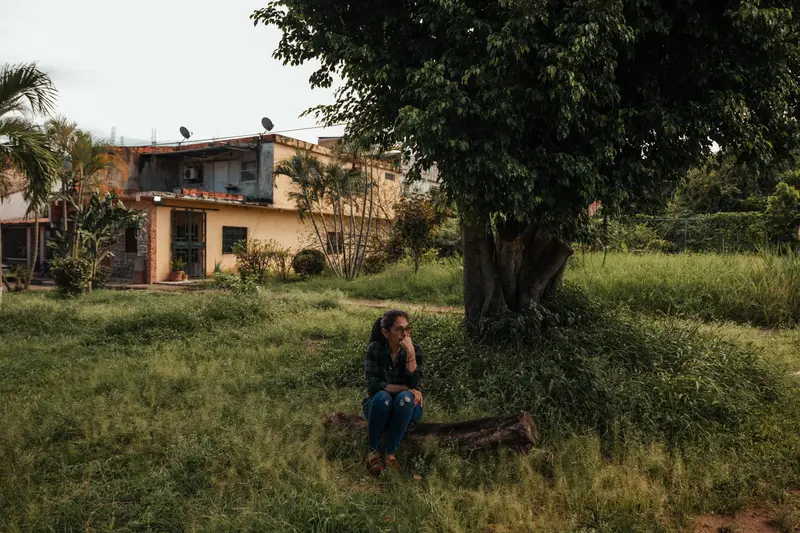 Eine Frau in Brille und Jeans sitzt auf einem Baumstamm unter einem Baum in der Nähe eines Hauses.