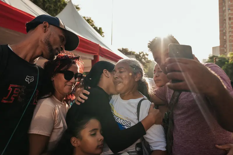 Sechs Personen, darunter ein Kind, drängen sich um eine Frau mit grauem Haar in einer Gruppenumarmung. Sie befinden sich im Freien neben einem Zelt, und der Mann weiter rechts hält ein Telefon für ein Selfie.