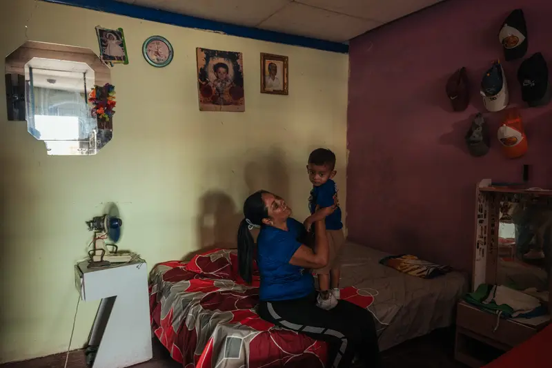 Eine Frau mit langen dunklen Haaren hält einen Jungen, der auf ihrem Schoß steht, als sie auf einem Bett sitzt. Ein Tischlüfter ist neben ihnen in Bewegung.
