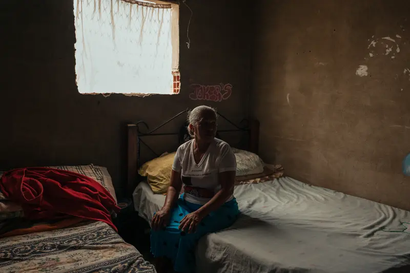 Eine Frau mit weißem Haar sitzt auf einem Bett in einem dunklen Raum. Die Wände sind schmucklos, ein weiteres Bett drängt sich in der Nähe dessen, auf dem sie sich befindet.
