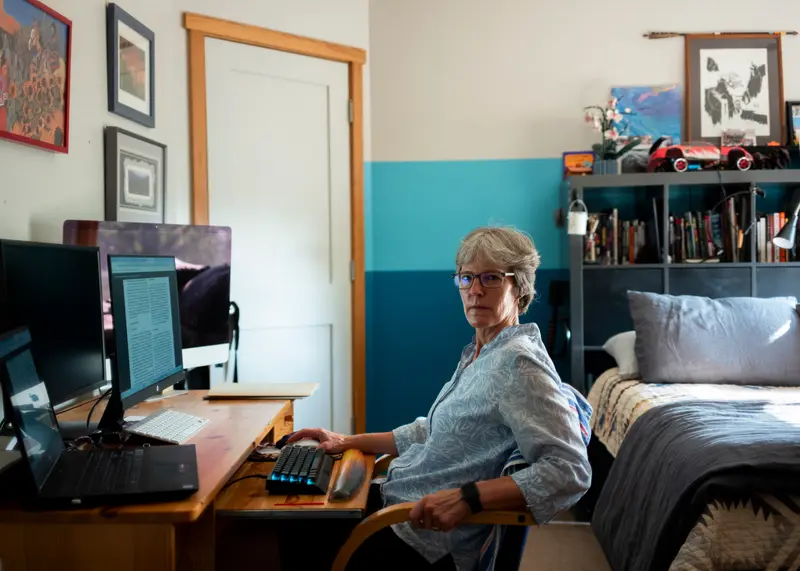 McCanlies sits at a wooden desk with multiple monitors and keyboards. Behind her is a made bed and a bookcase, with framed pictures hanging on the walls.