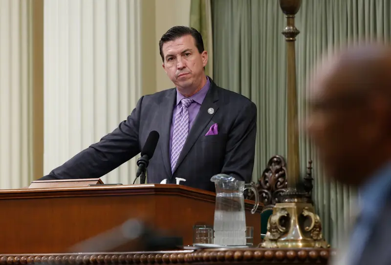 A man wearing a grey suit, purple tie, purple shirt and purple pocket square stands at a podium with curtains behind him.