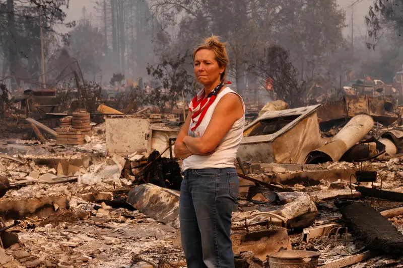 A woman wearing a U.S. flag bandana around her neck, jeans and a white tank top stands with her arms folded, surrounded by burnt trees and building debris.