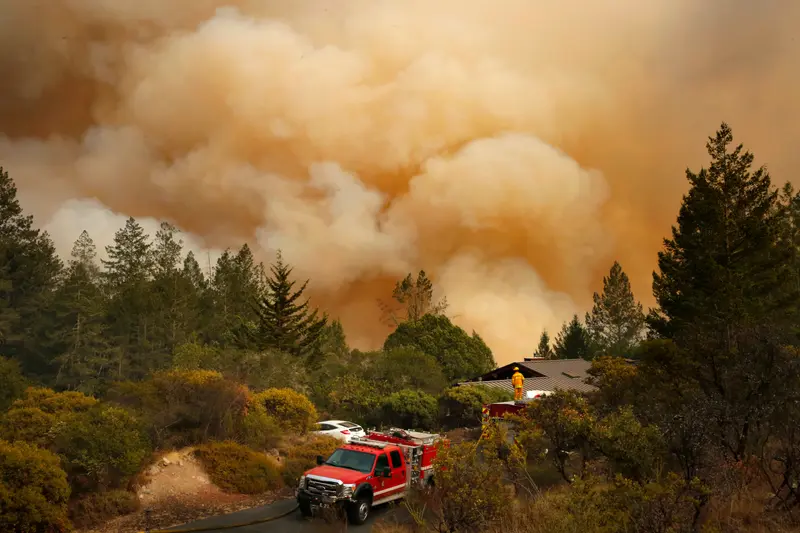 Large orange clouds fill the sky behind conifer trees, a firetruck and a lone firefighter in the distance.