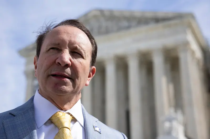 Close-up of a man from the chest up, wearing a blue suit and a yellow tie in front of a blurred courthouse.