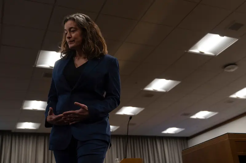 A woman in a blue suit stands looking down at her hands in a dim room.