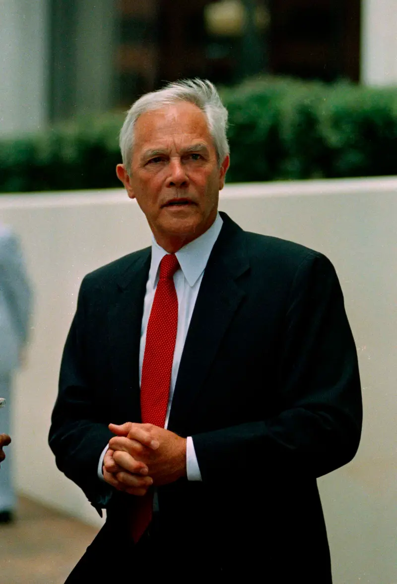 A man with white hair and wearing a blue suit and red tie clasps his hands in front of his body. He is standing outside in front of a white wall and a green hedge.