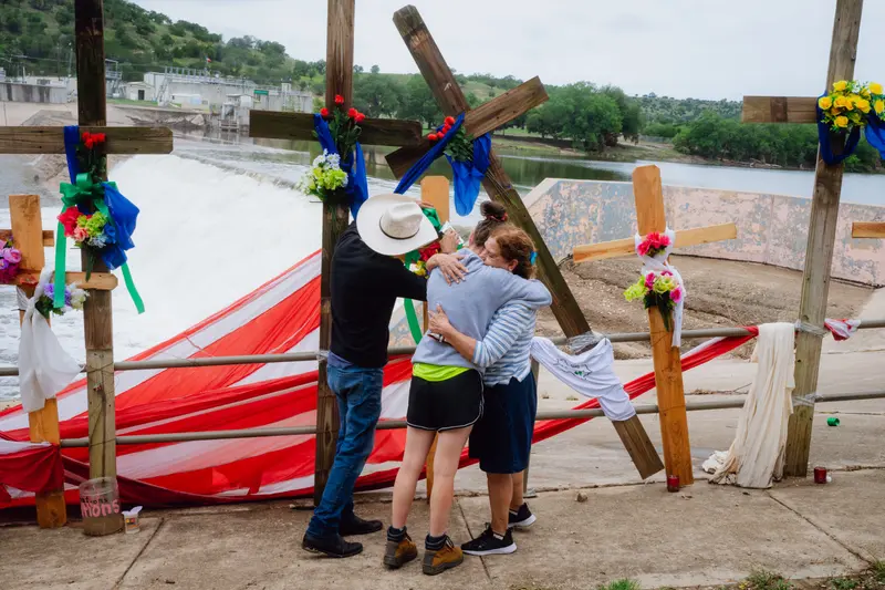 Three people face away from the camera, and two of them hug each other beside a row of wooden crosses with bouquets of flowers. They’re standing in front of a river dam.