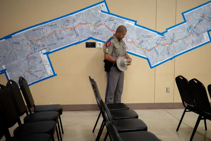 A law enforcement officer bows his head and holds a brimmed hat in his hand. He’s standing alone in a room filled with rows of empty black chairs. Behind him is a map of a river with handwritten notes, fixed to the wall with blue tape.