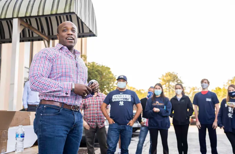 A Black man in a plaid shirt and jeans stands outside in front of a row of casually dressed people wearing face masks.