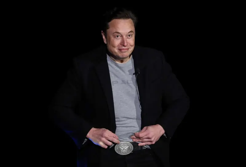 Musk, wearing a black blazer and a gray SpaceX T-shirt, lifts the hem of his shirt to show a large silver belt buckle featuring a five-pointed star with Tesla’s stylized “T” logo in the middle.
