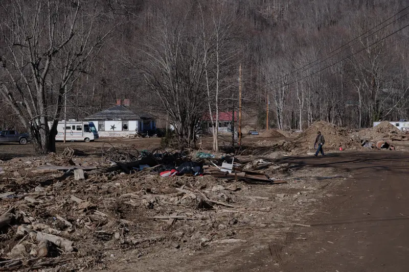 A man walks down a dirt road through a landscape of wooden debris and destruction.