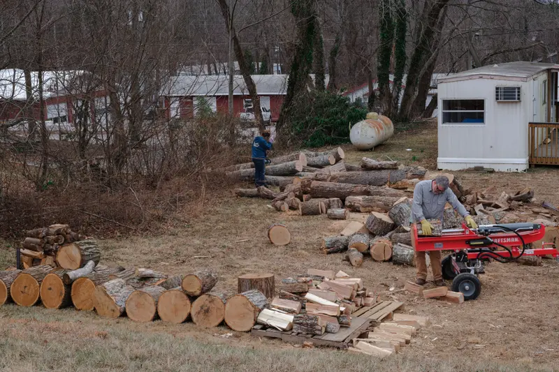 A man holds a section of wood on top of a red log-splitter. Behind him lies stacks of logs.