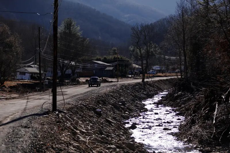 A car travels along a damaged road beside a wooded creek. Mountains and conifer trees appear in the distance.